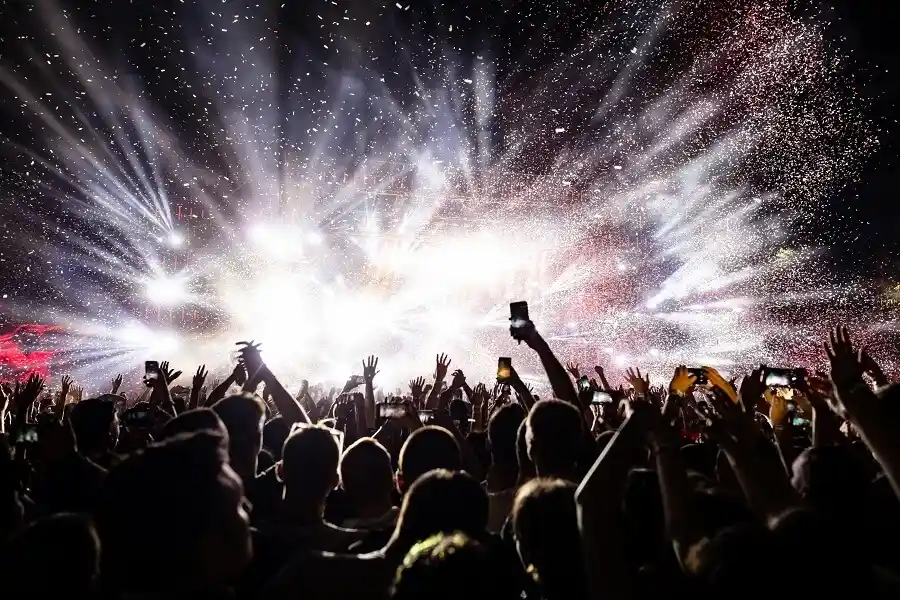 A cheering concert crowd with raised hands and smartphones beneath dramatic, bursting stage lights and falling confetti.
