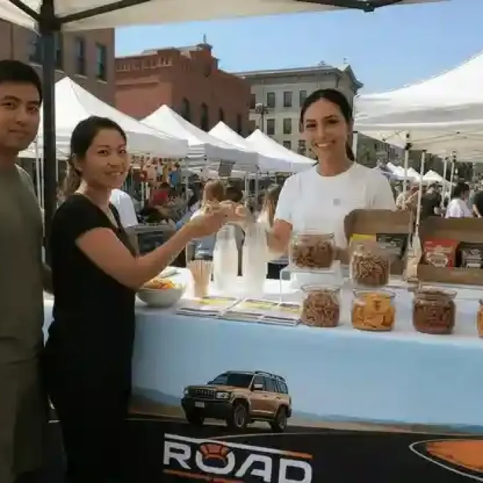A smiling vendor in a white t-shirt hands a sample across a display table to a customer at a sunny outdoor market under a canopy tent.